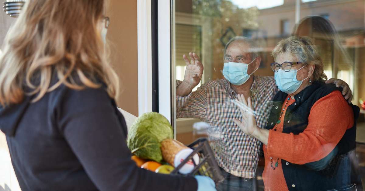 Couple in their home wearing masks, waving to family or friend delivering groceries
